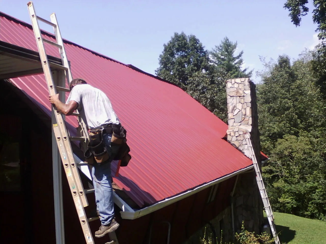 Roofer on a ladder beside a red metal roof and stone chimney — Soffit & Fascia Repair by Roofing Brothers in Randleman